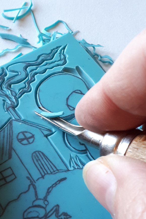 Main gravant une plaque de linoléum bleue, motif de maison, lors d’un atelier au musée