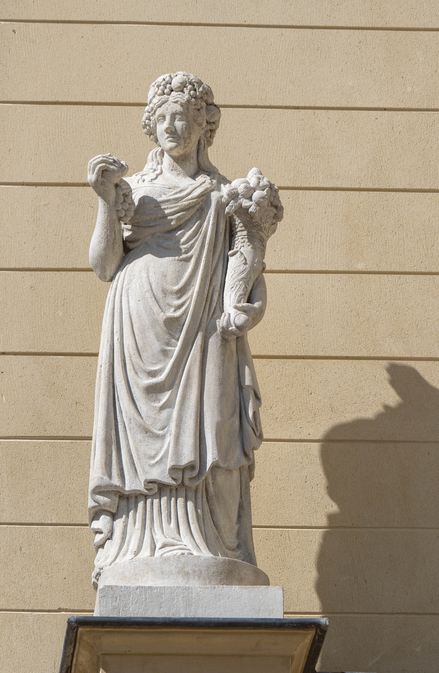 Statue en pied drapée, tenant une corne d’abondance dans une main et une grappe de raisin dans l’autre