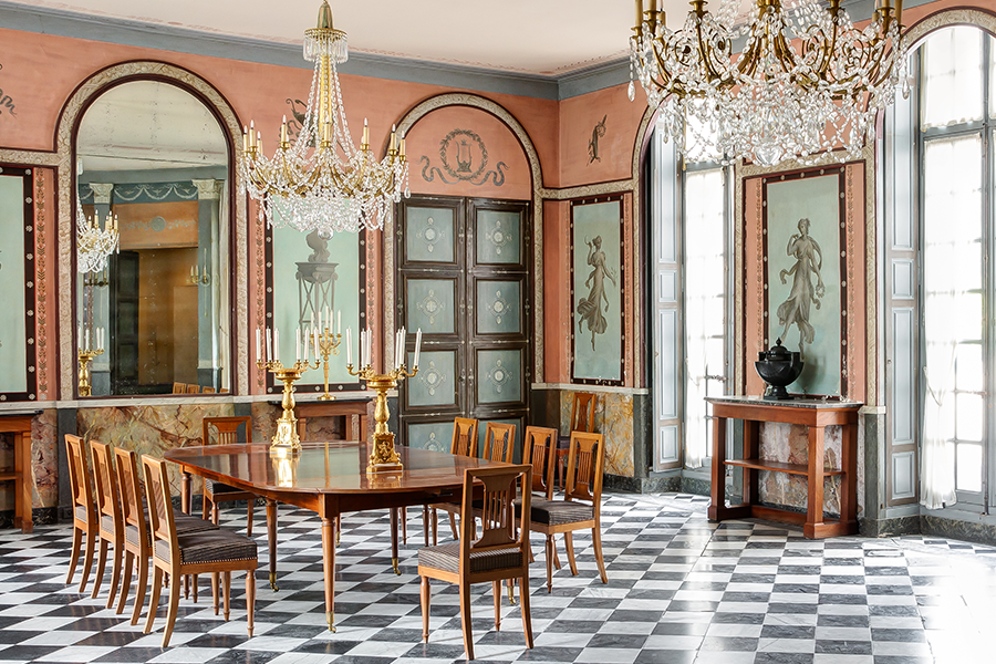 Longue table entourée de chaises, sous des lustres en cristal, dans un décor de fresques pompéiennes